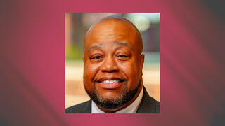 Corporate headshot of Dr. Derrick Jordan smiling in a dark suit and tie with a blurred office background.