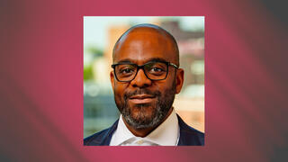 Corporate portrait of Jamaal K. Livan wearing glasses and a navy suit against a soft cityscape background.