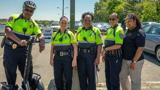 Five campus police officers and staff pose together outdoors in a parking lot near an "Event Parking" sign.