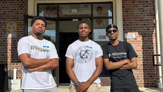 Three young men pose with crossed arms outside a university building.