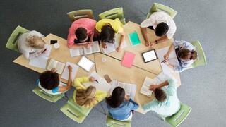 An overhead view of a group of students sitting around modular wooden tables studying together with books, notebooks, and digital tablets.