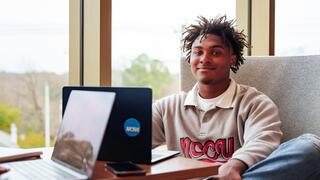 A smiling student wearing an NCCU sweatshirt sits at a desk with open laptops near a large window.
