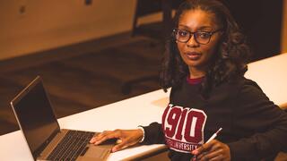 A woman wearing glasses and a dark university sweatshirt holding a pen sits at a desk working on an open laptop.  