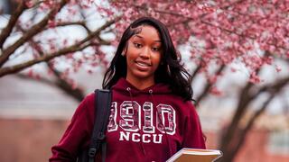 A smiling student wearing a maroon NCCU hoodie holds a notebook in front of blooming pink cherry blossom trees.