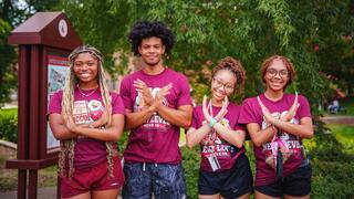 Four students wearing matching maroon NCCU move-in day t-shirts pose together outdoors while doing the eagle sign