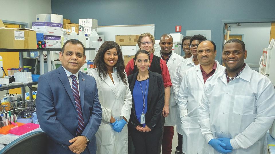 Eight people, several in white coats, stand in a laboratory in a semi-circle.