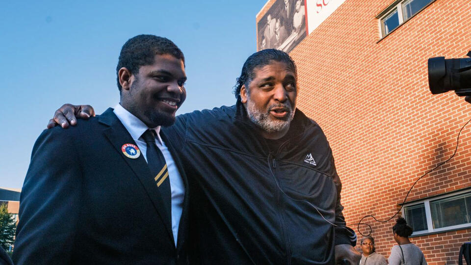 Two men stand outside a brick building, one in a suit with his arm around the other man, who is wearing a black jacket.
