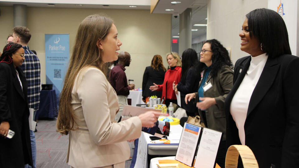 A professional woman with long hair in a light-colored suit stands and talks to another professional woman at a career fair, with other attendees and booths visible in the background.