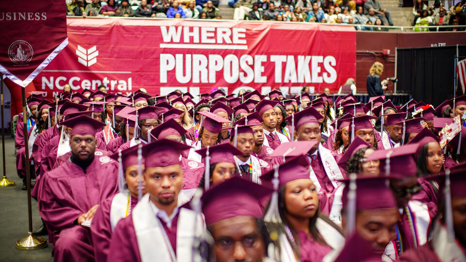 NCCU graduates in maroon caps and gowns seated at commencement ceremony beneath “Where Purpose Takes Flight” banner.