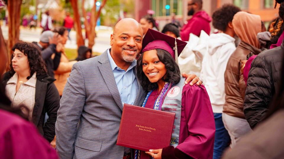 Graduate in cap and gown poses with family member, holding degree during NCCU commencement celebration.