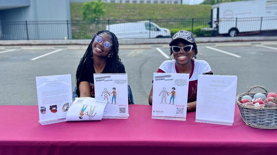 Two smiling women sit behind a pink table displaying wellness campaign materials and a basket of apples.