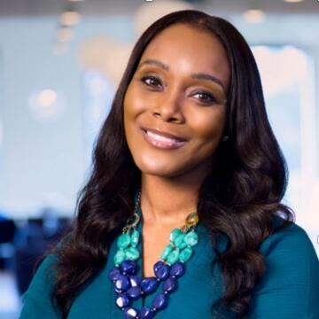 Professional headshot of Jerraé “Raé” Williams smiling in a teal top with a layered blue and green statement necklace, standing in a bright office setting.