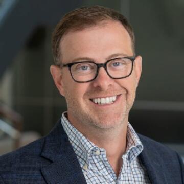 Professional headshot of Josh Parker smiling, wearing glasses, a blue blazer, and a checked shirt, posed in a modern office setting.