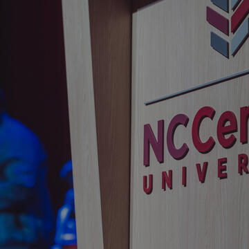  The image shows the "NCCentral University" logo on a wooden surface, with a person playing a saxophone in the blurred background.