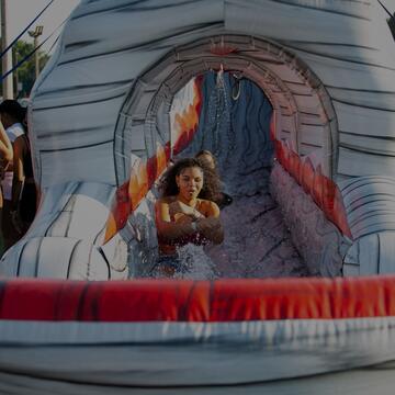 Students sliding down a water slide at an outdoor campus event, with splashes of water and others waiting in line nearby.