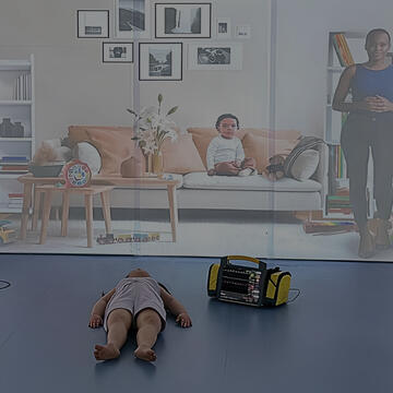 A child laying on the floor in front of a screen displaying a woman and a baby in a living room, with a yellow device beside the child.