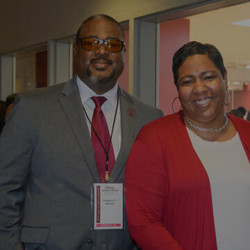 A man and woman smiling and standing together indoors. The man wears a gray suit with a red tie and name badge, and the woman wears a red cardigan over a white top.