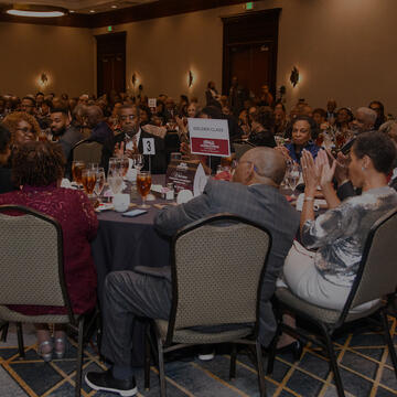 A large group of people dressed in formal attire sit at round tables during a banquet event, with a sign reading “Golden Class” displayed at one table.