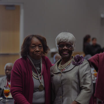 Two older women smile while posing together at a formal luncheon. One wears a maroon sweater with a medal, and the other wears a silver suit with pearls and a purple flower accessory.