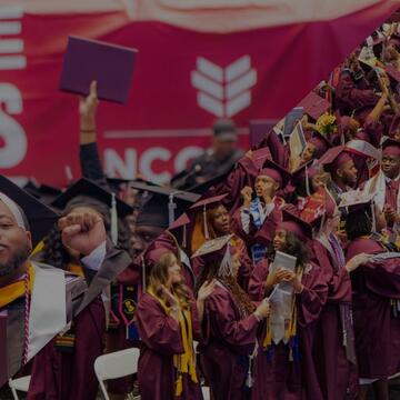 NCCU graduates celebrating in McDougald-McLendon Arena, wearing maroon caps and gowns, holding diplomas, and cheering during commencement.