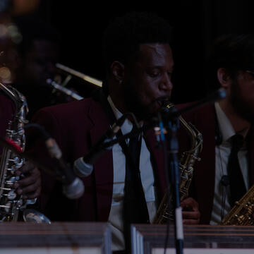 Jazz musicians perform on stage wearing maroon blazers and black ties, playing saxophones and brass instruments during a live concert.