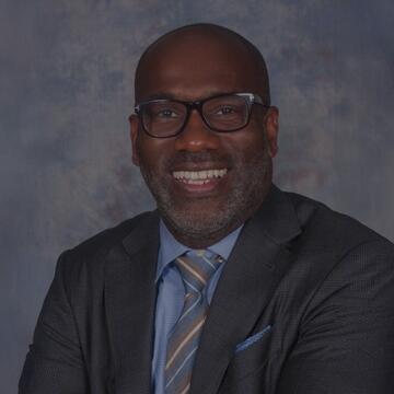 Professional headshot of Joseph Karl Grant smiling, wearing glasses and a suit with tie against a neutral studio background.