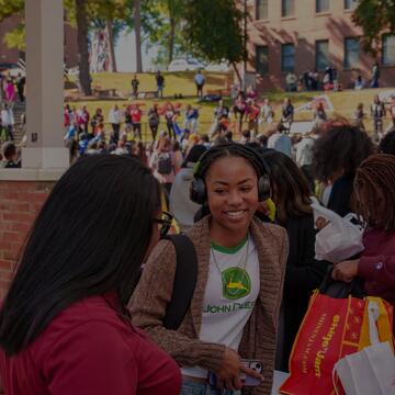 A smiling woman wearing over-ear headphones and a John Deere shirt stands outdoors amidst a large crowd at a college campus event.
