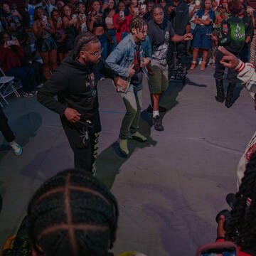 A group of young men perform in the center of an open arena floor surrounded by a crowd recording with their phones.