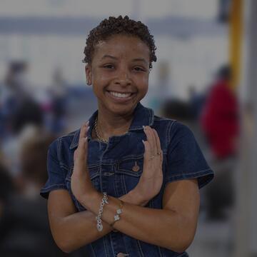 A smiling young woman in a denim top does the eagle sign against a blurred, busy indoor background.