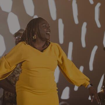 A smiling woman in a long-sleeved bright yellow dress sings onstage in front of a choir with a patterned backdrop illuminated behind them.