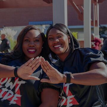 Two smiling women pose together making hand signs in front of a covered outdoor courtyard.