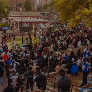 A large crowd gathers around a campus courtyard and reflecting pool surrounded by autumn trees during a university event.