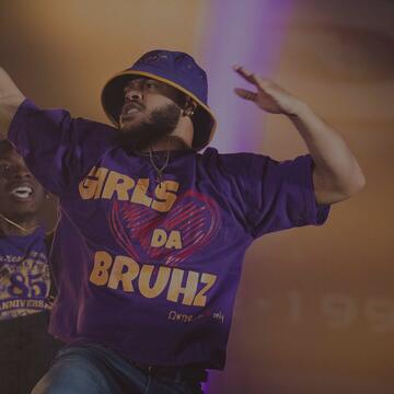 A man wearing a purple bucket hat and a t-shirt reading "GIRLS LOVE DA BRUHZ" performs dynamically on stage alongside other group members